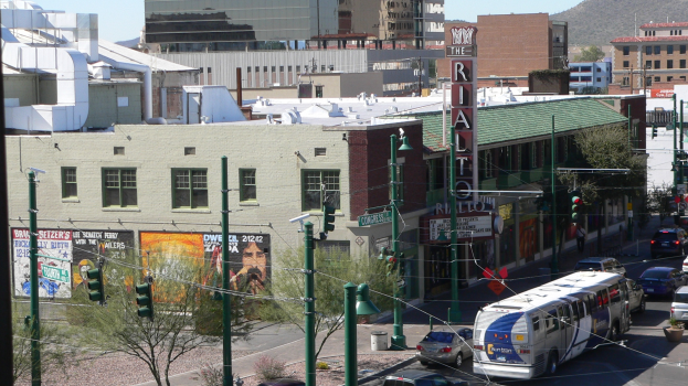 Eine belebte Stadtstraße mit verschiedenen Fahrzeugen, darunter ein Bus, Autos und ein Landfahrzeug, umgeben von Pfählen, Laternen, Schildern, Bäumen, Gebäuden, einem Hügel und einem Himmel.