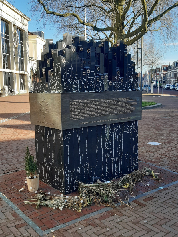 Denkmal in einem Stadtplatz für Holocaust-Opfer, mit einem Baum davor, einem Topfgewächs, verstreuten trockenen Blättern und Gebäuden, Fahrzeugen und Bäumen im Hintergrund.