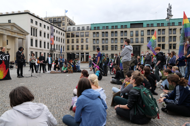 Eine Gruppe von Menschen, die auf dem Boden sitzen, vor einer Menge mit Fahnen und Spruchbändern, mit einer Person, die in ein Mikrofon spricht, einer Statue und Gebäuden im Hintergrund bei einer Homo-Protest in Berlin.