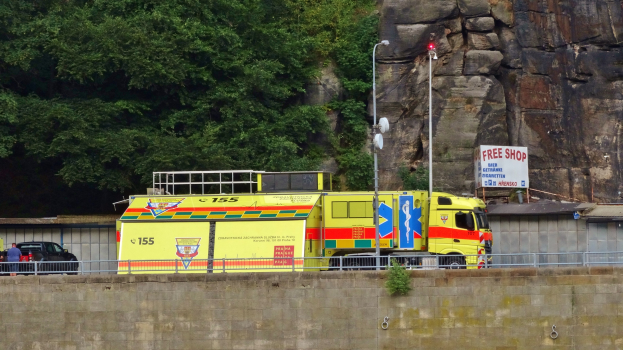 Ambulanz auf der Seite einer Straße neben einer Klippe geparkt, mit verschiedenen Gegenständen wie einer Wand, einem Gelönder, Pfosten, Lichtern und Schildern mit Text im Vordergrund und Bäumen und einem Auto im Hintergrund.