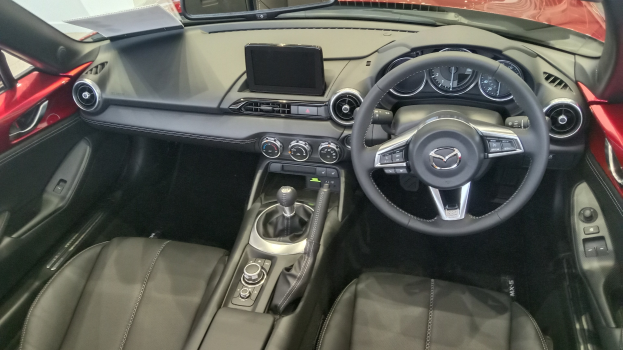 Innenansicht eines roten Mazda MX-5 Miata RF mit schwarzen Ledersitzen, Lenkrad, Schaltknüppel, Knöpfen, Bildschirm, Spiegel und anderen Innenraumkomponenten.