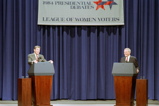 Zwei Männer in formeller Kleidung stehen an Podien mit Mikrofonen und Gläsern vor einem blauen Vorhang und einem Banner mit Text und Sternen, diskutieren die Debatten von 1984.
