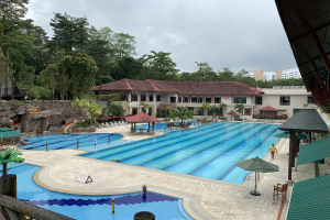 Großes Schwimmbad im Ritz-Carlton, Kota Kinabalu, mit Menschen, Sonnenschirmen, Liegestühlen, Tischen, Grünpflanzen, Felsen und Gebäuden, einschließlich eines Schuppens auf der rechten Seite und Himmel im Hintergrund.