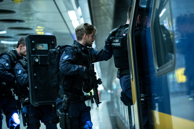 Eine Gruppe von Polizisten steht neben einem Zug auf der rechten Seite des Bildes, wobei einer der Beamten eine Waffe hält und an der Decke Lampen befestigt sind.