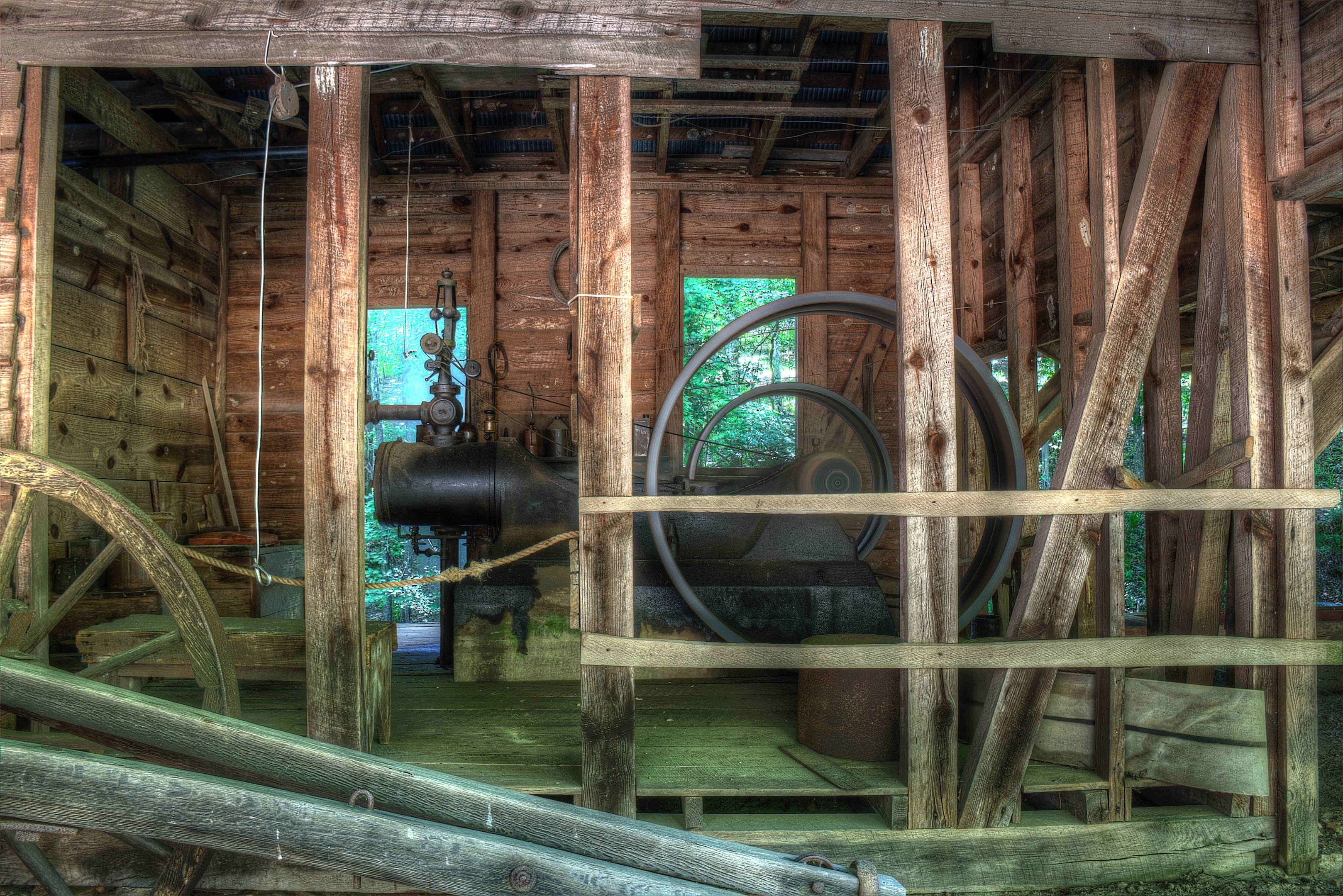 Altes Holzgebäude mit großer Industrieanlage, gestützt von Holzpfählen und einem Rad, mit Bäumen im Hintergrund.