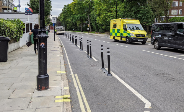 Ambulanz auf einer Straße neben einem Bürgersteig mit fahrenden Fahrzeugen, einem Fußgänger, einem Müllcontainer, Versorgungsmasten, Schildern, Bäumen, Gebäuden und einem bewölkten Himmel.