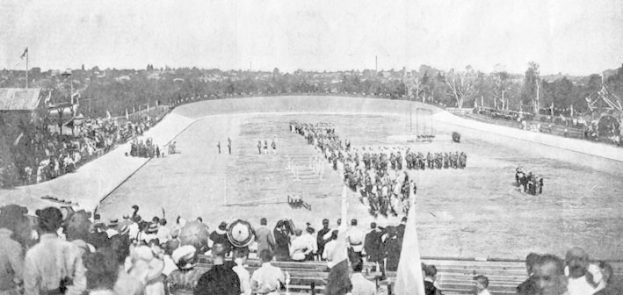 Altes Schwarz-Weiß-Foto von Zuschauern bei einem Pferderennen im Stadion, mit Menschen auf Bänken und Stehplätzen, einige halten Fahnen und andere reiten Pferde, vor einem Hintergrund aus Bäumen und einem klaren Himmel.
