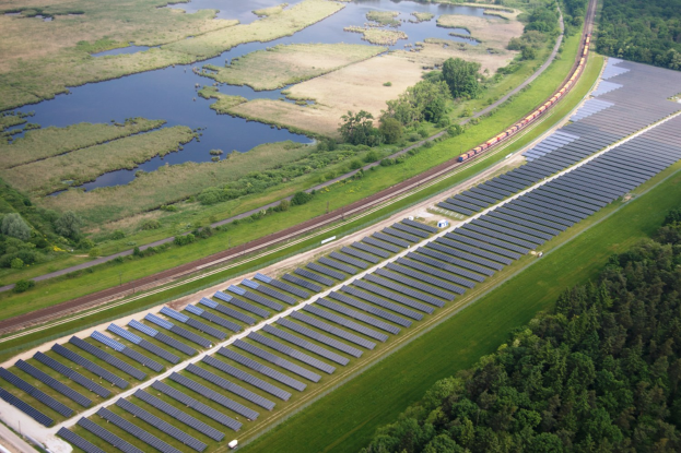 Luftaufnahme eines Solarparks mit Solarpanelen in einem Feld, umgeben von Bäumen, Gras, Wasser und einer nahen Bahnlinie.