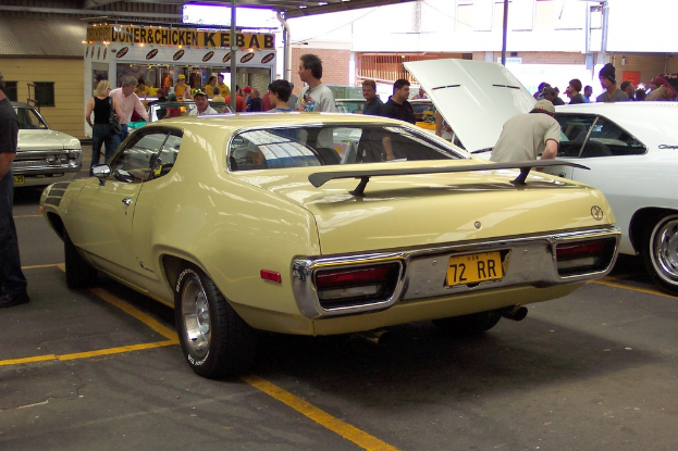 Ein Oldtimer steht auf einer Straße mit Menschen in der Nähe, ein Geschäft in der Mitte und Gebäuden im Hintergrund.