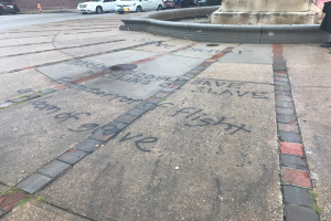 Geweg mit "Black Lives Matter"-Graffiti in großen Buchstaben, umgeben von einem Gebäude, Fahrzeugen, Menschen, Bäumen, Pfählen und einer Wand.