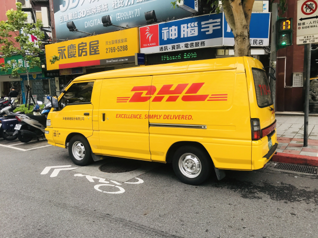 Gelber DHL-Lieferwagen auf der Straße neben Gebäuden, Bäumen, Schildern, Polen, Ampeln und einem Fußgänger auf dem Bürgersteig.