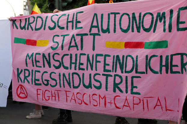 Eine Gruppe von Menschen hält eine pinkfarbene Fahne mit der Aufschrift "Kurdische Autonomie Statt Menschenfeindlicher Kriegsindustrie Fight Fascism Capital" auf einer Straße mit Ampeln, Flaggen, Bäumen, Gebäuden und einem klaren blauen Himmel im Hintergrund.