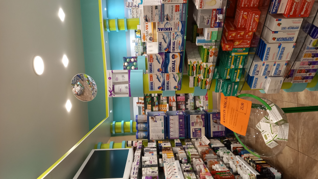 Eine Apotheken-Theke mit verschiedenen Medikamenten in Schachteln und Flaschen auf Regalen, einem Spiegel an der Wand, Deckenleuchten und einem Glastisch mit Papieren und Gegenständen darauf, der Boden ist unten zu sehen.