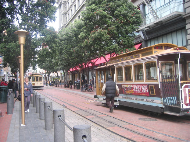 Eisenbahnschienen verlaufen durch eine Straße mit Zügen, auf denen Text zu sehen ist, einer Person in der Nähe, kleinen Pfählen mit Seilen auf der linken Seite, Straßenlaternen, Bäumen, vielen Menschen im Hintergrund, einem Müllcontainer auf der linken Seite und Gebäuden mit Leitern auf der rechten Seite.