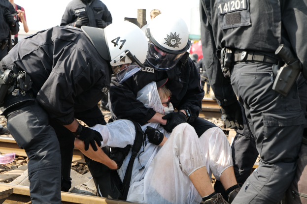 Eine Gruppe von Polizisten in Einsatzausrüstung verhaftet einen Mann in einem weißen Kleid auf einem Gleis, mit anderen im Hintergrund sichtbar.