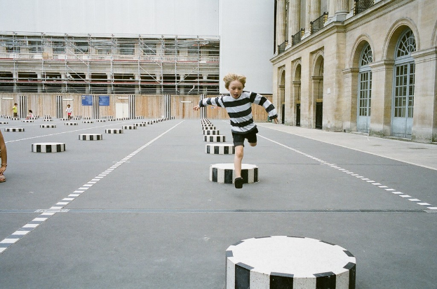 Ein Spielbereich mit vier Kindern, eines läuft auf dem Boden mit schwarzen und weißen Betonstufen, umgeben von einem Schulgebäude.