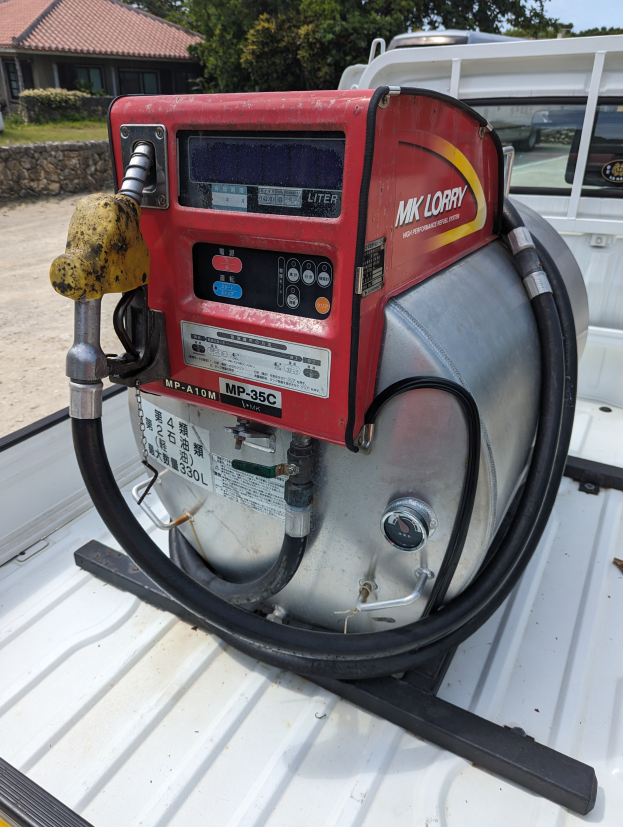 Ein Lastwagen mit einer Tankröhre im Heck, umgeben von Bäumen, einem Haus und einem klaren blauen Himmel, ausgestattet mit einem Motor und einem Kraftstoffspender.