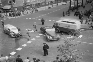 Schwarze und weiße Szene eines Autounfalls an einer Straßenkreuzung mit mehreren Fahrzeugen, darunter ein Van, und einer Gruppe von Zuschauern; Gebäude, Bäume, Strommasten und Texttafeln sind im Hintergrund zu sehen.