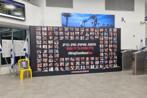 Eine große Wand zeigt zahlreiche Fotos, einen gelben Stuhl, israelische Regierungsflaggen auf Stangen, eine Tafel mit dem Text "Bringt sie jetzt nach Hause", einen montierten Bildschirm, Deckenleuchten und andere Gegenstände.