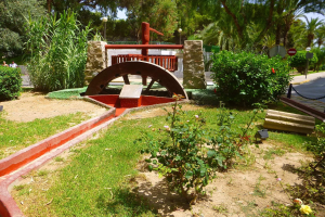 Ein Minigolf-Platz in einem Park mit Grünfläche, einer Straße mit Fußgängern und Schildern sowie Bäumen unter einem klaren blauen Himmel.