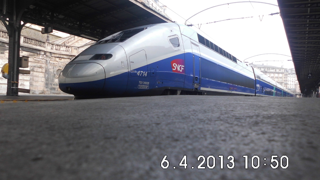 Ein Zug ist in der Mitte des Bildes mit einem Bahnsteig und Text unten, Gebäuden und einem Dach links und Gebäuden, Dächern, Kabeln und Himmel oben.