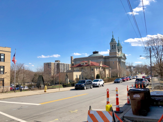 Stadtstraße mit parkenden Autos, Verkehrskegeln, Mülltonnen, einem Feuerhydranten, einem Zaun, Bäumen, Gebäuden mit Fenstern, einer Flagge an einem Mast und einem bewölktem Himmel.