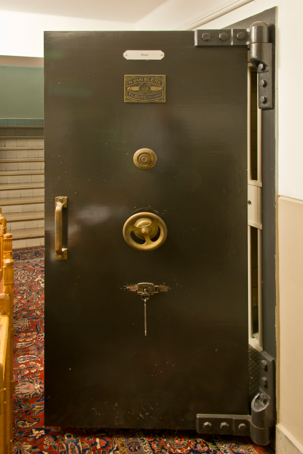 Eine große Metalltresortür mit einem Schloss und einem Schlüssel, umgeben von hölzernen Gegenständen auf der linken Seite, Treppe und Wand im Hintergrund und einer Decke darüber.
