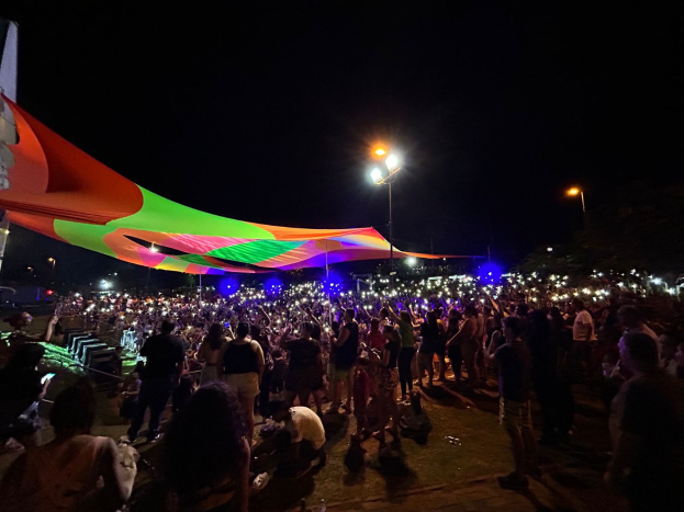 Eine große Menschenmenge versammelt sich unter bunten Lichtern nachts vor einem Zelt, mit einem Gebäude links und dem Himmel im Hintergrund während des 2018er Kunstfestivals.