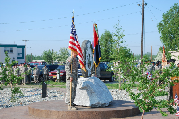 Zwei Militärangehörige stehen mit Fahnen auf beiden Seiten eines Gedenksteins, mit Pflanzen, Autos, anderen Militärangehörigen, Strommasten mit Kabeln und Gebäuden im Hintergrund.