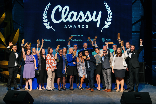 Eine Gruppe von Menschen auf einer Bühne, die strahlen und Preise halten, mit Lautsprechern unten und einem Hintergrundbild mit der Aufschrift "Classy Awards 2019."