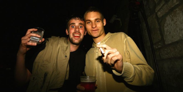 Zwei Männer stehen, jeder hält ein Glas in einer Hand und eine Zigarette in der anderen.