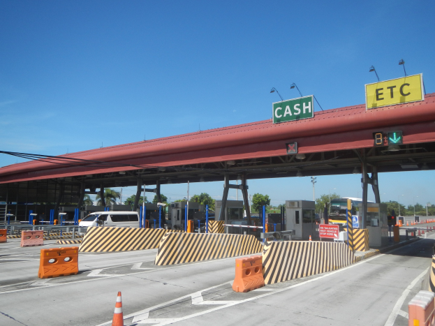 Eine Mautstelle mit einem "Bar"-Schild, Fahrzeugen, Schranken, Schildern, ├ffentlichen Stra├čenlaternen, Strommasten, Kabeln, B├Ąumen und einem bew├Âlktem Himmel im Hintergrund.