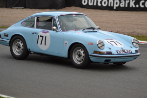 Ein blauer Porsche 911 GTS auf einer Rennstrecke mit einem Helmträger am Steuer, umgeben von Gras und einem schwarzen Banner mit Text im Hintergrund.