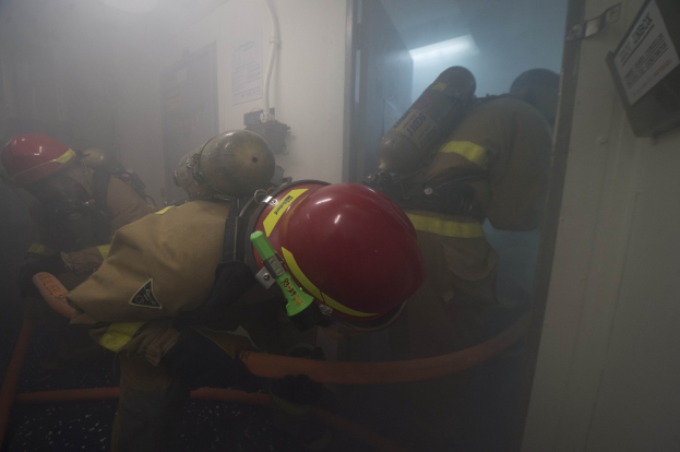 Feuerwehrleute in Schutzausrüstung betreten einen raucherfüllten Raum, mit Sauerstofftanks und Rohren sichtbar.