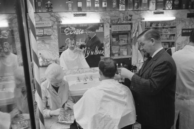 Schwarz-weißes Foto eines Mannes, der einer Frau in einem Friseursalon die Haare schneidet, mit einem Waschbecken, Spiegel, Regalen und einer Glastür im Hintergrund.