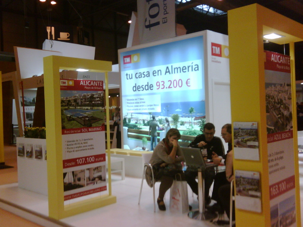 Eine Gruppe von Menschen sitzt um einen Tisch und schaut auf einen Laptop, mit verschiedenen Werbeplakaten daneben.