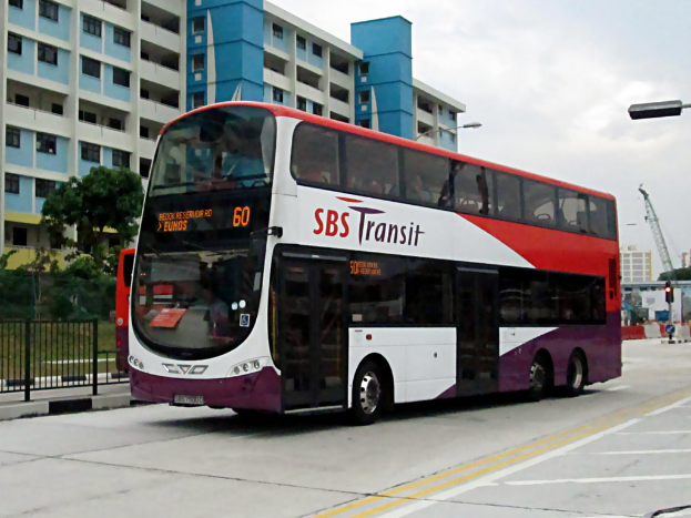 Ein Elektrobus ist in der Mitte des Bildes mit einem Gebäude im Hintergrund, Bäumen vor dem Gebäude, einem Zaun auf der rechten Seite des Fußwegs und Himmel oben.