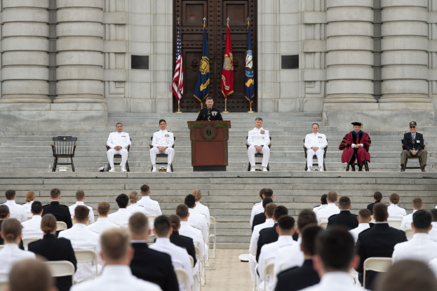 Eine Gruppe von Menschen sitzt auf Stühlen vor einem Gebäude mit Säulen und einer Tür, mit einem Podium mit Mikrofon in der Mitte, Flaggen mit Stangen im Hintergrund und einem leeren Stuhl auf der linken Seite, was auf eine Abschlussfeier hinweist.