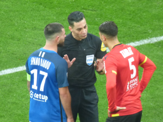 Schiedsrichter spricht mit zwei Männern auf einem Fußballfeld, einer trägt eine schwarze Hose und ein schwarzes Hemd, der andere ein blaues und rotes T-Shirt mit Text, beide tragen eine Brille.