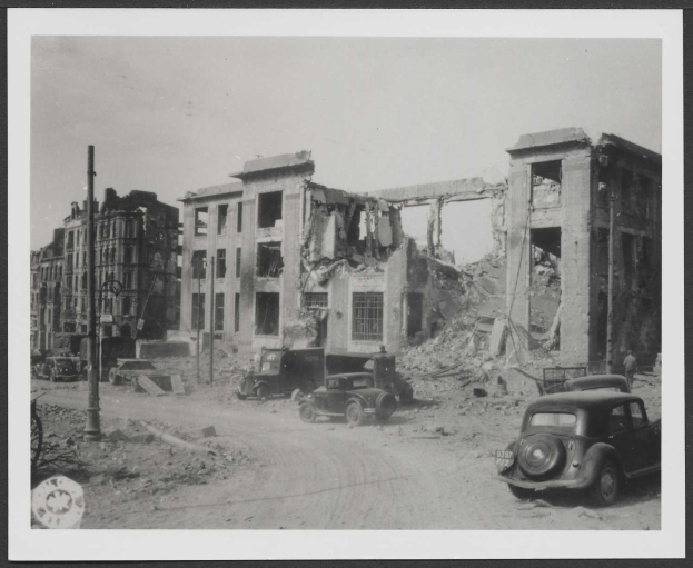 Schwarz-weißes Bild eines zerstörten Gebäudes in Berlin mit parkenden Autos davor, wahrscheinlich aus einem Bombenarchiv.