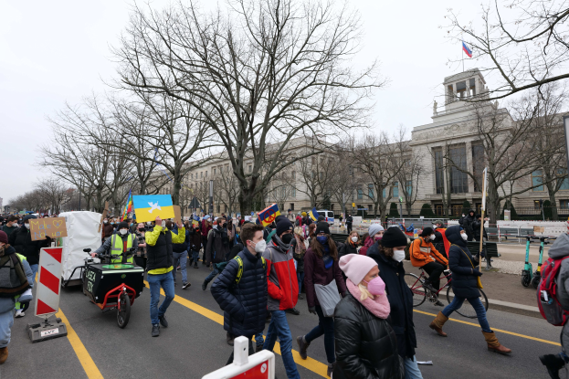 Eine große Protestdemo mit Menschen, die eine Straße in Washington, D.C. entlanggehen, einige halten Schilder und andere fahren Fahrräder, mit Bäumen und einem klaren blauen Himmel im Hintergrund.
