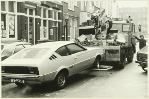 Ein Schwarz-Weiß-Foto eines Autos, das von einem Abschleppwagen auf einer Straße mit anderen Fahrzeugen, einer Person in der Nähe und Gebäuden mit Fenstern im Hintergrund abgeschleppt wird.