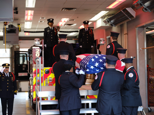 Gruppe uniformierter Männer, die einen Sarg tragen, der mit einer amerikanischen Flagge bedeckt ist, vor einem Feuerwehrauto und Deckenlampen im Hintergrund.
