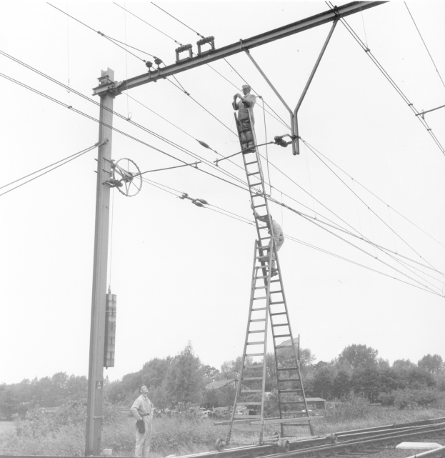 Zwei M"anner auf Leitern bei der Arbeit an einer Stromleitung mit einem Pfahl im Vordergrund, B"umen und Himmel im Hintergrund und Eisenbahnschienen unten.