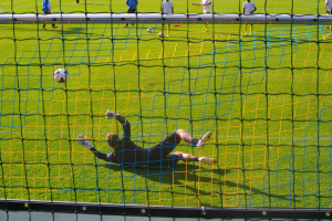 Ein Fussballspieler taucht ab, um einen Schuss zu halten, mit einem Tor im Vordergrund und einigen Menschen im Hintergrund, während mehrere Fussbälle auf dem Boden liegen.