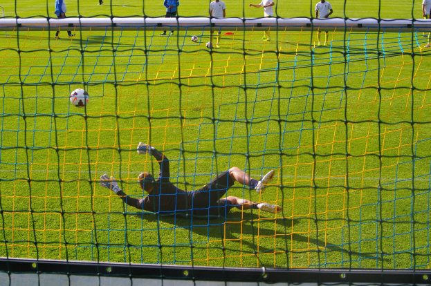Ein Fussballspieler taucht ab, um einen Schuss zu halten, mit einem Tor im Vordergrund und einigen Menschen im Hintergrund, während mehrere Fussbälle auf dem Boden liegen.