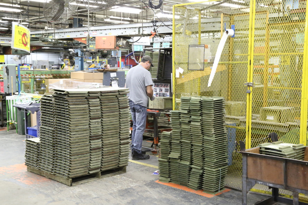 Ein Mann in einer Mütze und T-Shirt steht in einer Fabrik, arbeitet in der Nähe von Maschinen auf dem Boden und an der Decke, mit Lüftern und Lampen darüber.