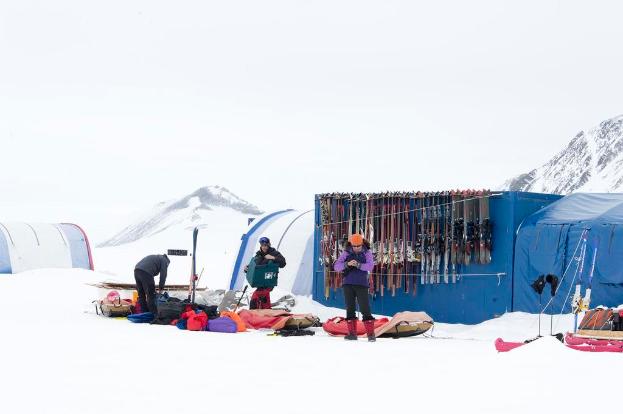 Drei Personen stehen auf einer verschneiten Landschaft mit Taschen drumherum, Zelten mit Skiern dahinter und schneebedeckten Hügeln im Hintergrund unter einem klaren Himmel.
