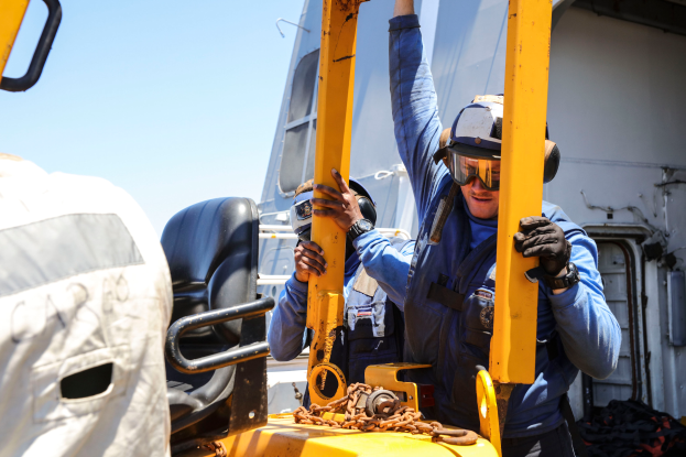 Zwei Männer in blauen Uniformen und Helmen arbeiten an einem gelben Stapler, tragen Handschuhe und Schutzbrillen, wobei einer ein Metallobjekt hält, ein Fahrzeug und der Himmel im Hintergrund.