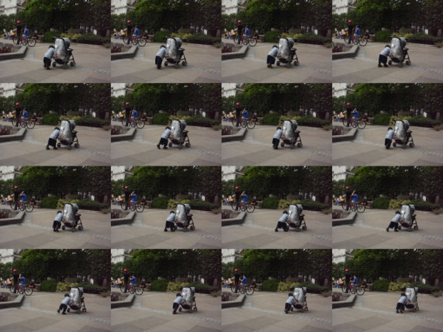 Ein Kind geht auf einer Straße und zieht einen Handwagen, mit zwei Personen, die Fahrräder halten, links, Bäumen im Hintergrund und einem Gebäude links.